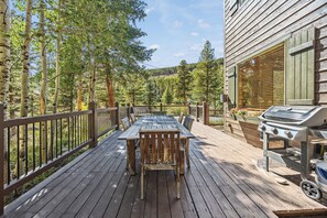 Una terraza de madera con mesa y sillas, una parrilla y vistas a un área boscosa.