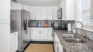 A modern kitchen with white cabinets, a stainless steel refrigerator, a granite countertop, and a window with blinds. - Steps to Beach! |Sleeps 11!| Summer Ready! (Panama City)