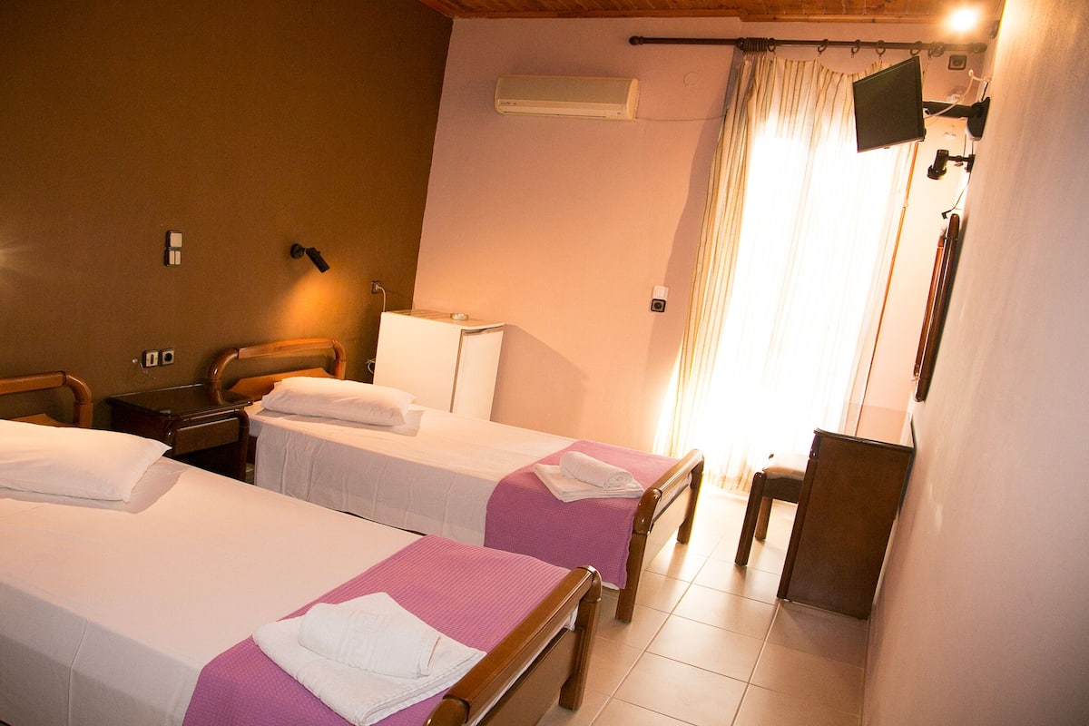 Une chambre d’hôtel avec deux lits simples, un petit bureau, une chaise, un téléviseur fixé au mur et une fenêtre avec des rideaux.