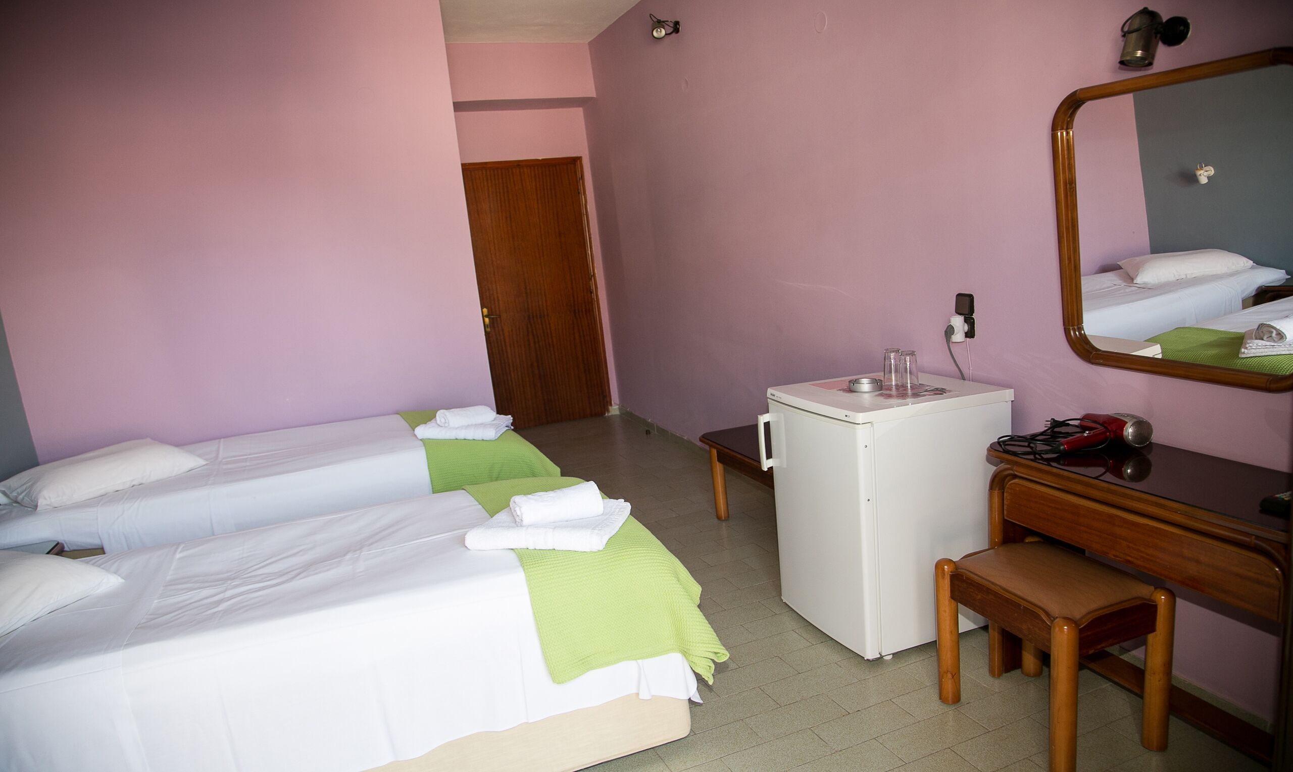 A hotel room with two single beds, a small desk with a mirror, a compact refrigerator, and a wooden chair.