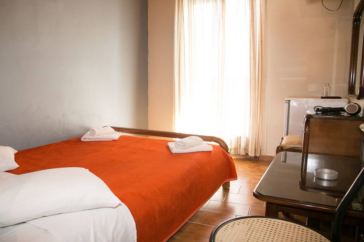 Une chambre d’hôtel avec deux lits, l’un recouvert d’une couvre-lit orange et l’autre de draps blancs. On y trouve une petite table avec une chaise, un miroir et une radio.