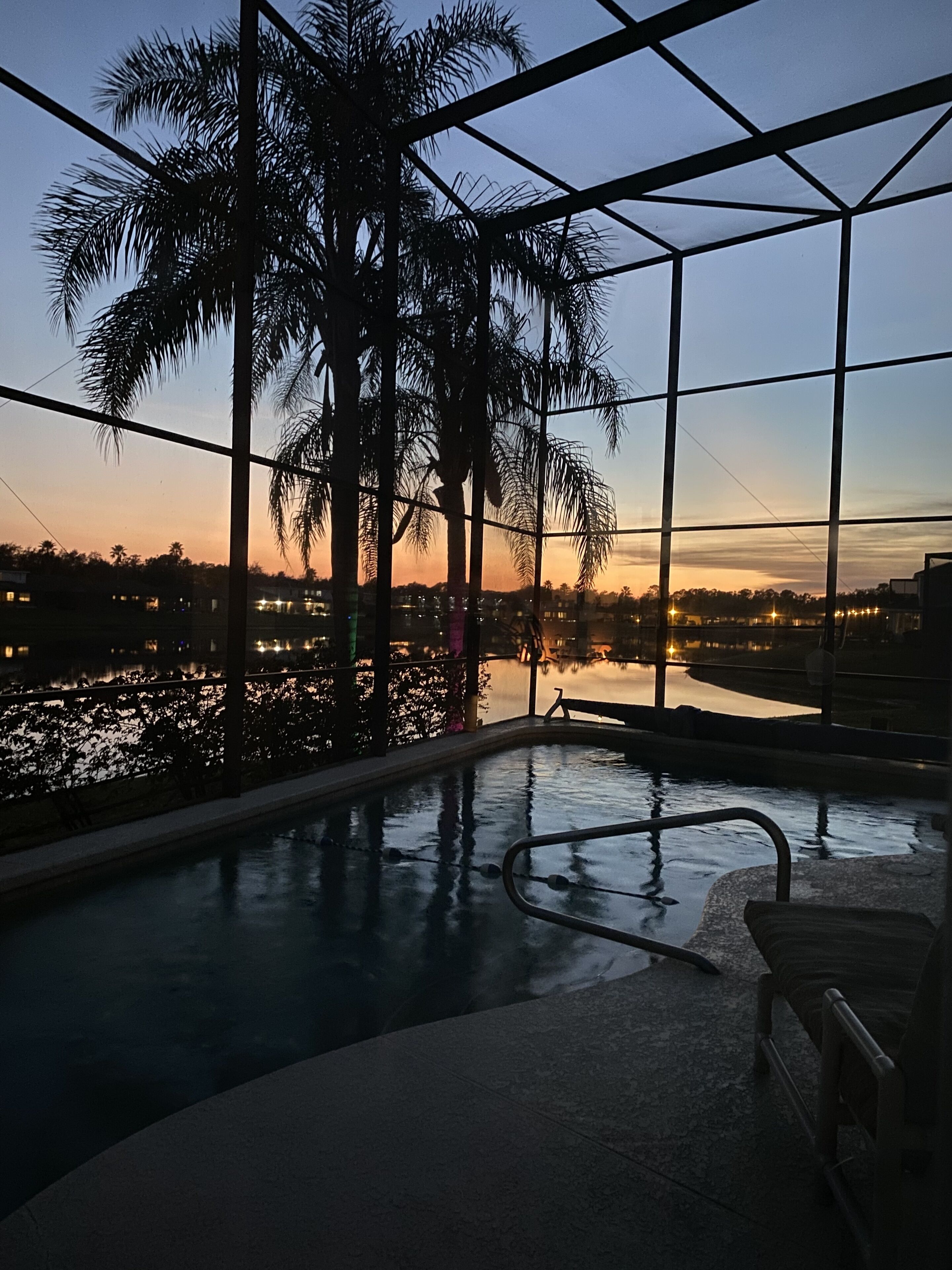 Une piscine intérieure offrant une vue sur un coucher de soleil se reflétant sur une étendue d’eau, le tout entouré de palmiers et protégé par une structure en verre.
