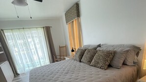 Een groot bed met een gestuufd hoofdeinde, een plafondventilator en uitzicht op de buitenwereld door een raam met dunne gordijnen.