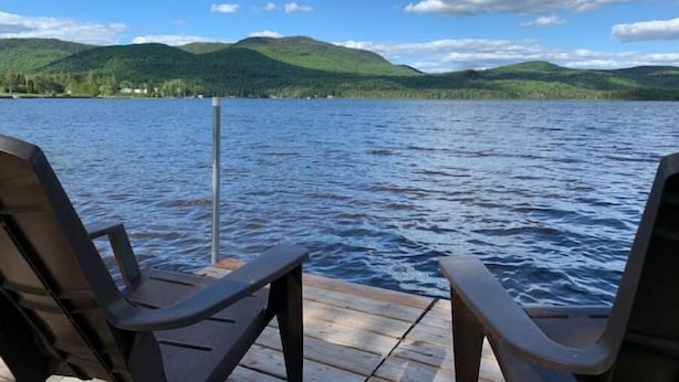 Une jetée en bois avec deux chaises, donnant sur un lac paisible, entourée de collines verdoyantes sous un ciel dégagé.