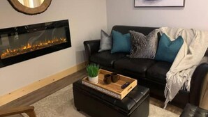 Un salon doté d’une cheminée, d’un canapé en cuir noir agrémenté de coussins turquoise, d’une table basse avec un plateau et d’un tableau représentant un paysage, accroché au mur.