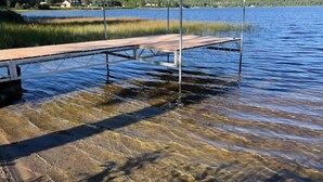 Une jetée en bois s’étendant dans un lac aux eaux claires, avec en arrière-plan des collines et quelques bâtiments.