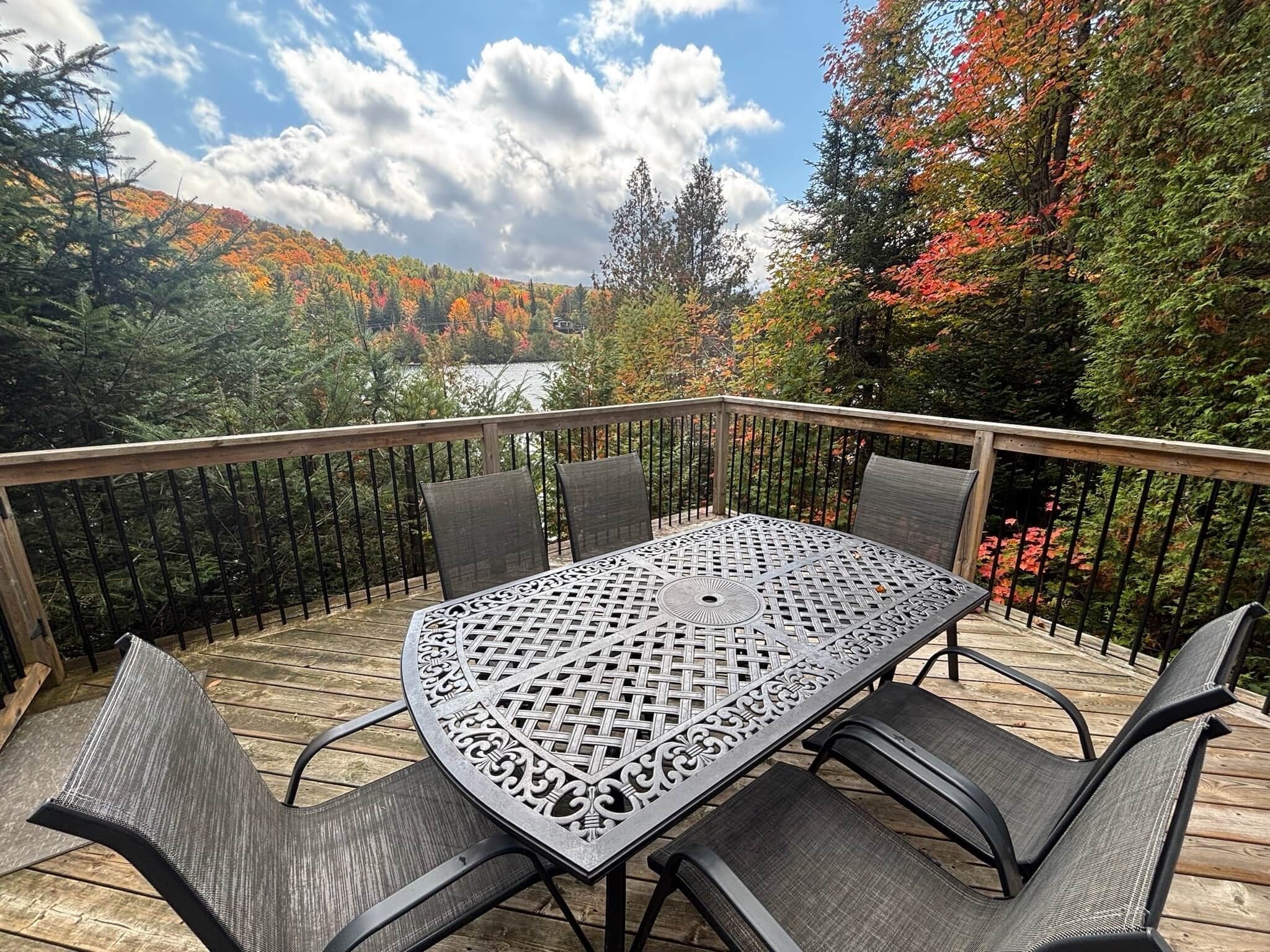 A deck with a table and chairs, surrounded by a wooden railing, overlooking a forested area with a lake.