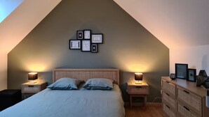 A bedroom with a sloped ceiling, a bed with pillows, two bedside tables with lamps, a dresser, and framed pictures on the wall.