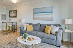 A living room with a grey sofa, a coffee table, a painting of the ocean, a lamp, and a small table with a plant. - Ocean Views + Resort Pool! N Topsail Beach Condo (North Topsail Beach)