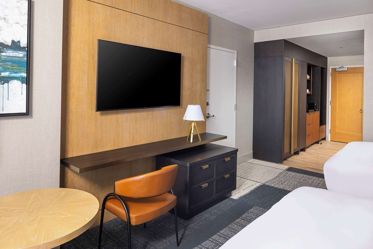A hotel room with a large flat-screen TV mounted on the wall, a wooden desk with a lamp, a black dresser, a round wooden table, and a white bed.
