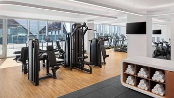 A modern gym with cardio machines, weight stations, and a large window offering a city view.
