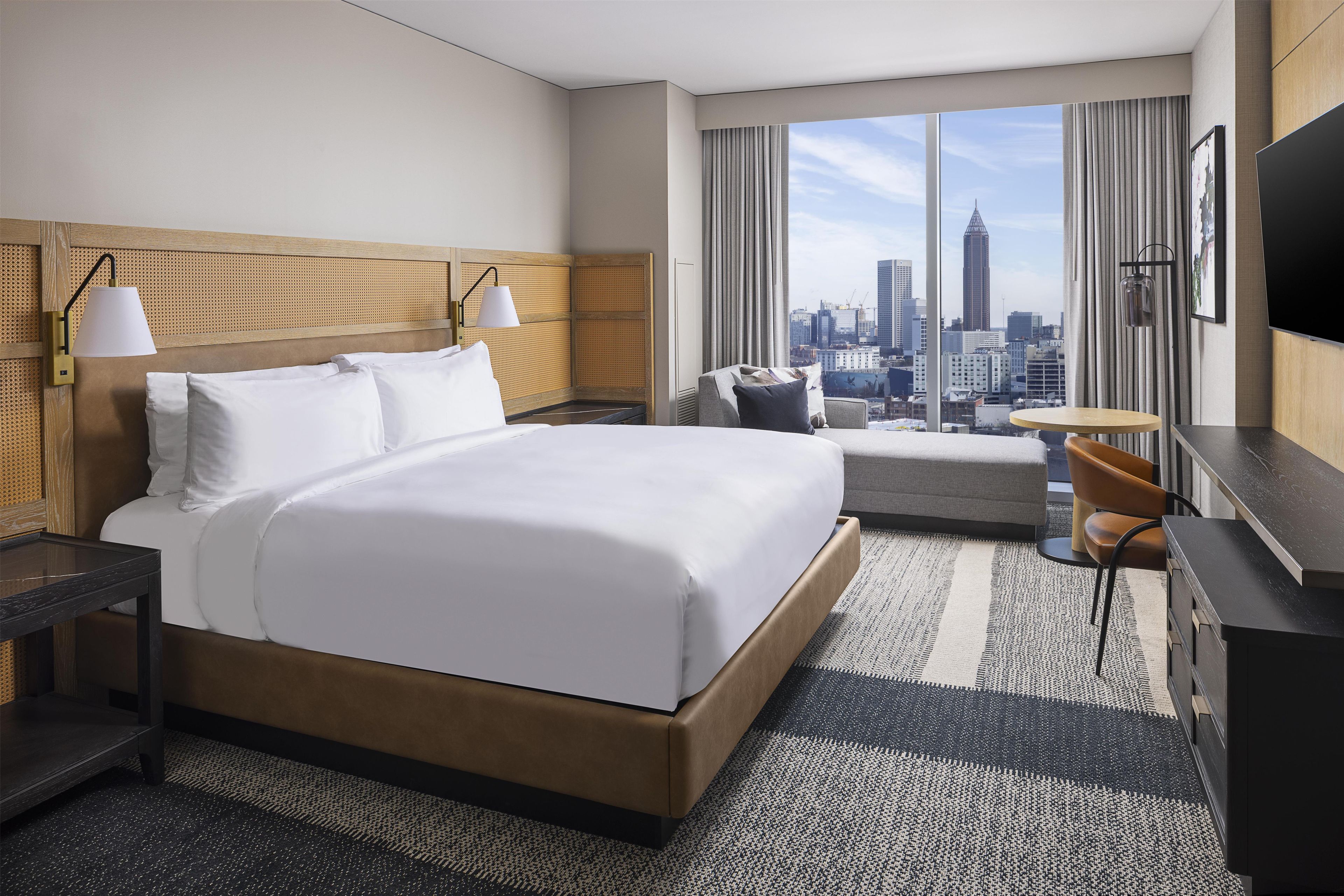 A hotel room with a large bed, a desk, a chair, a TV, and a view of a cityscape.