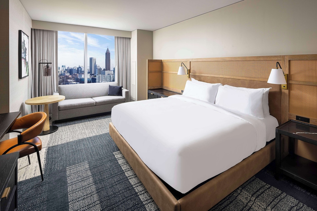 Quarto de hotel moderno com uma cama grande, um sofá, uma escrivaninha e vista para a cidade.
