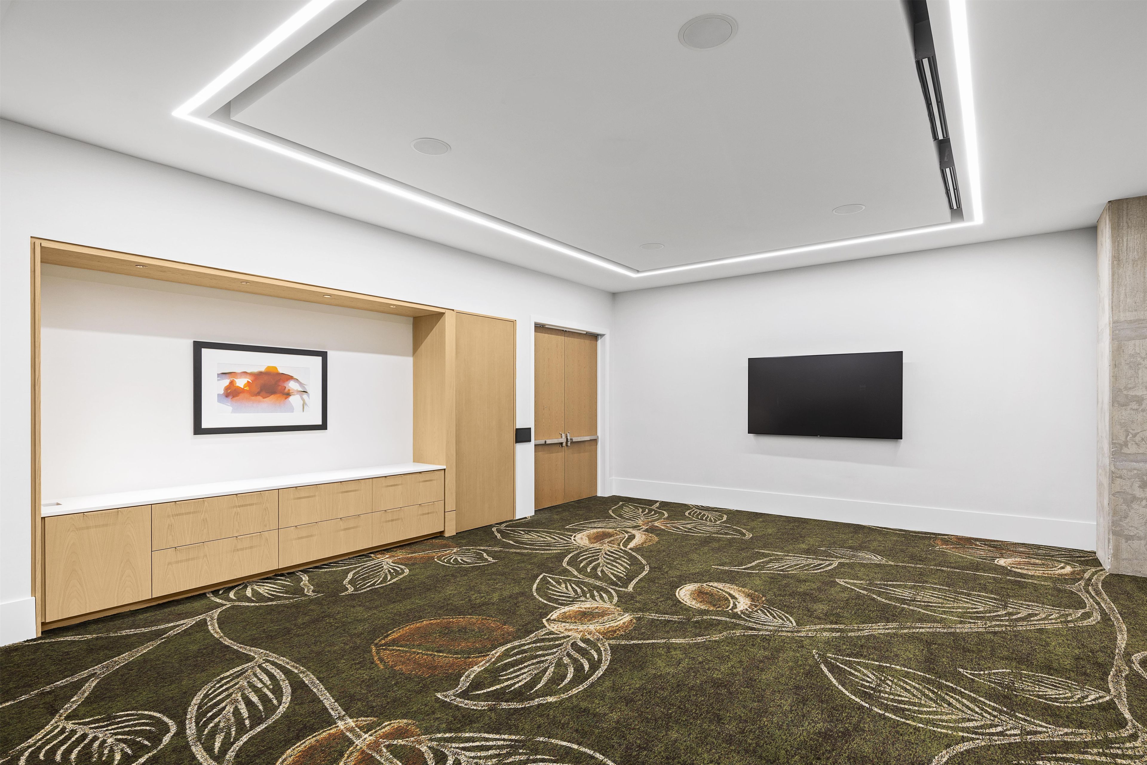 A room with a flat-screen TV mounted on the wall, a framed picture on the wall, a wooden cabinet with drawers, and a carpet with a leaf pattern.