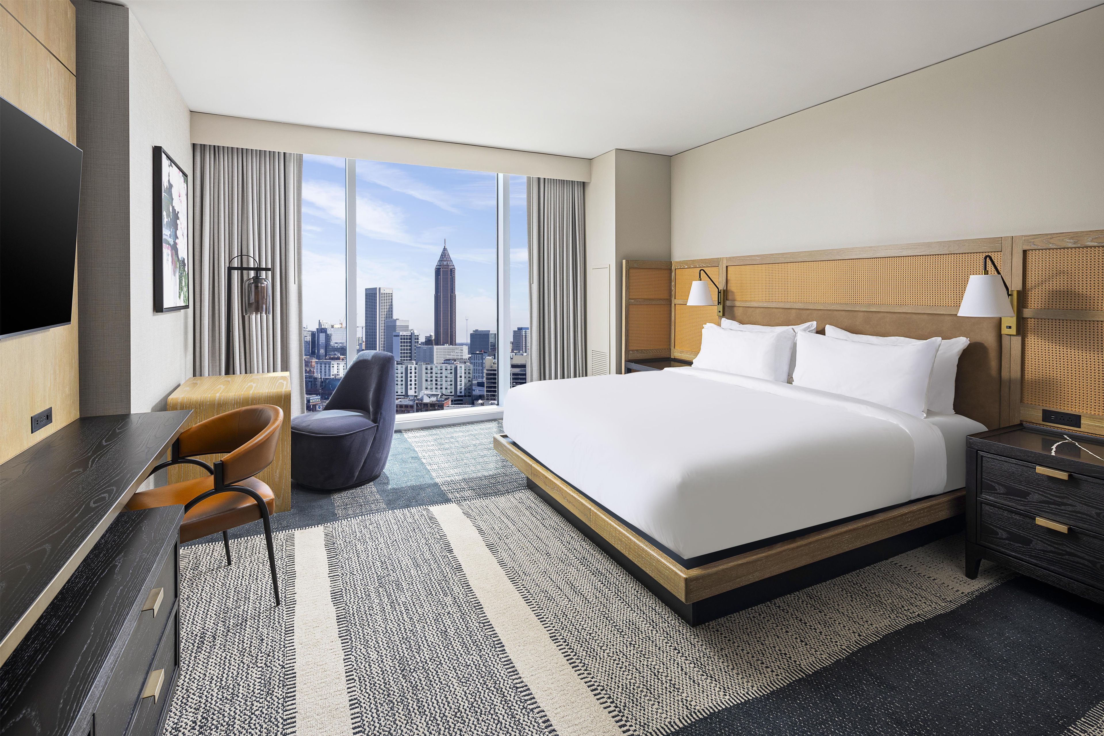 Habitación de hotel con una cama grande, un escritorio, una silla, una mesita de noche, una lámpara y vistas al horizonte de la ciudad.