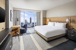Habitación de hotel con una cama grande, un escritorio, una silla, una mesita de noche, una lámpara y vistas al horizonte de la ciudad.