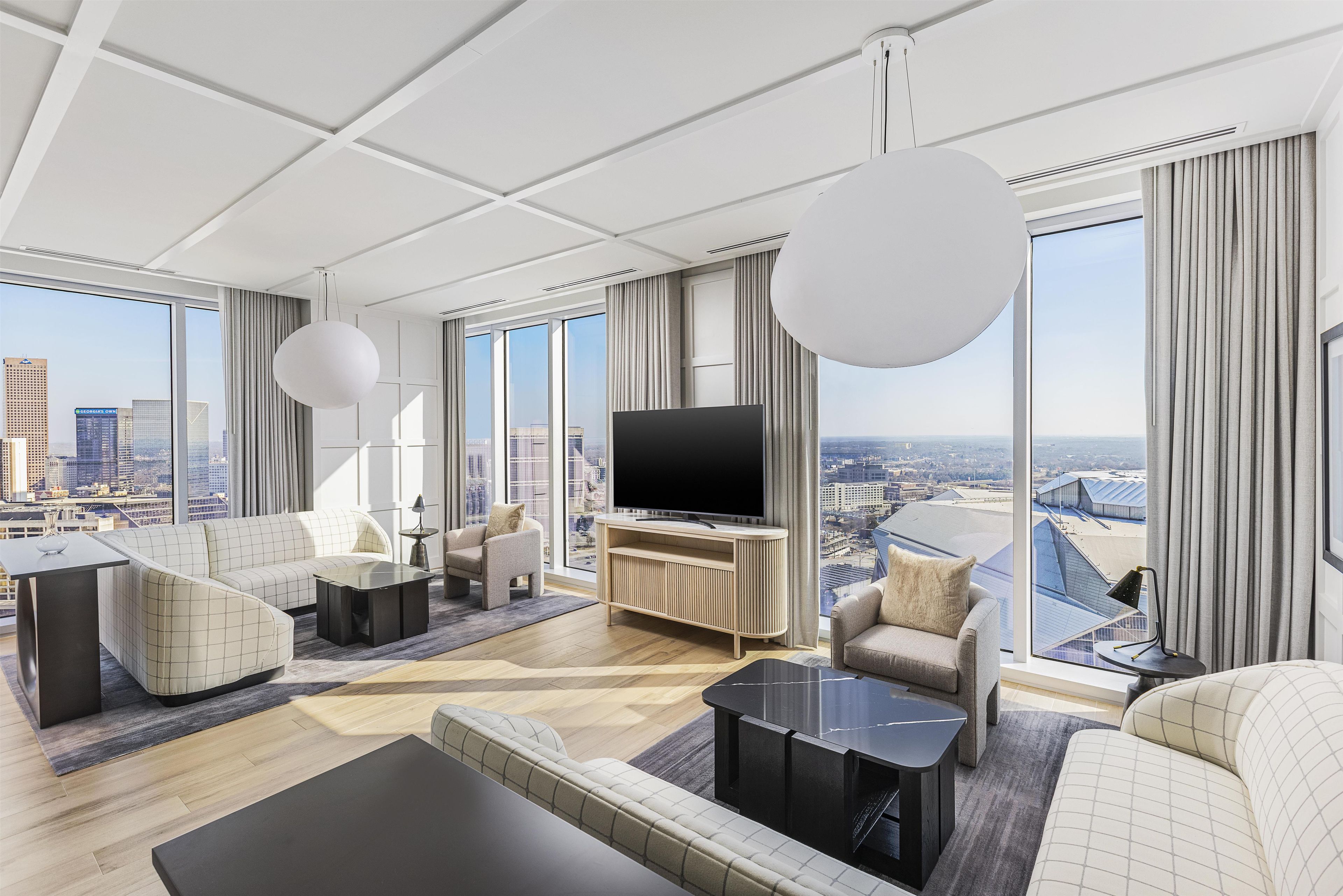 Habitación de hotel moderna con televisor grande, asientos cómodos, vista a la ciudad y un ventanal amplio.