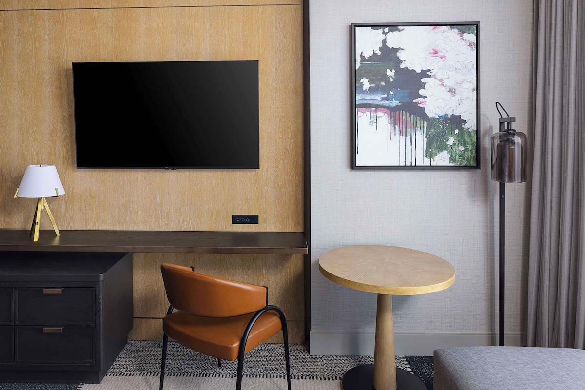 Quarto de hotel moderno com televisão de tela plana presa a uma parede de madeira, uma mesa de madeira redonda, uma cadeira de couro marrom, uma escrivaninha com gavetas, um abajur e uma obra de arte floral emoldurada.