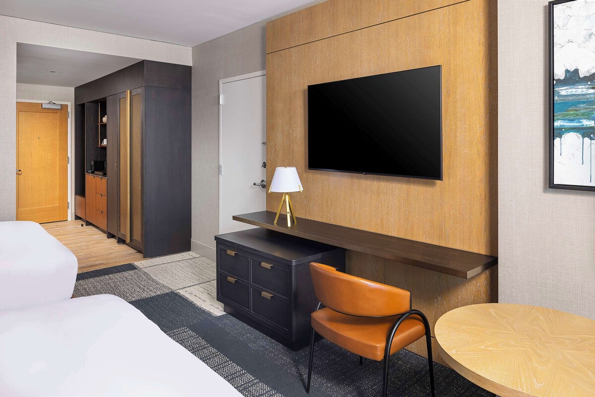 A hotel room with a bed, a desk with a lamp, a TV mounted on the wall, and a chair.