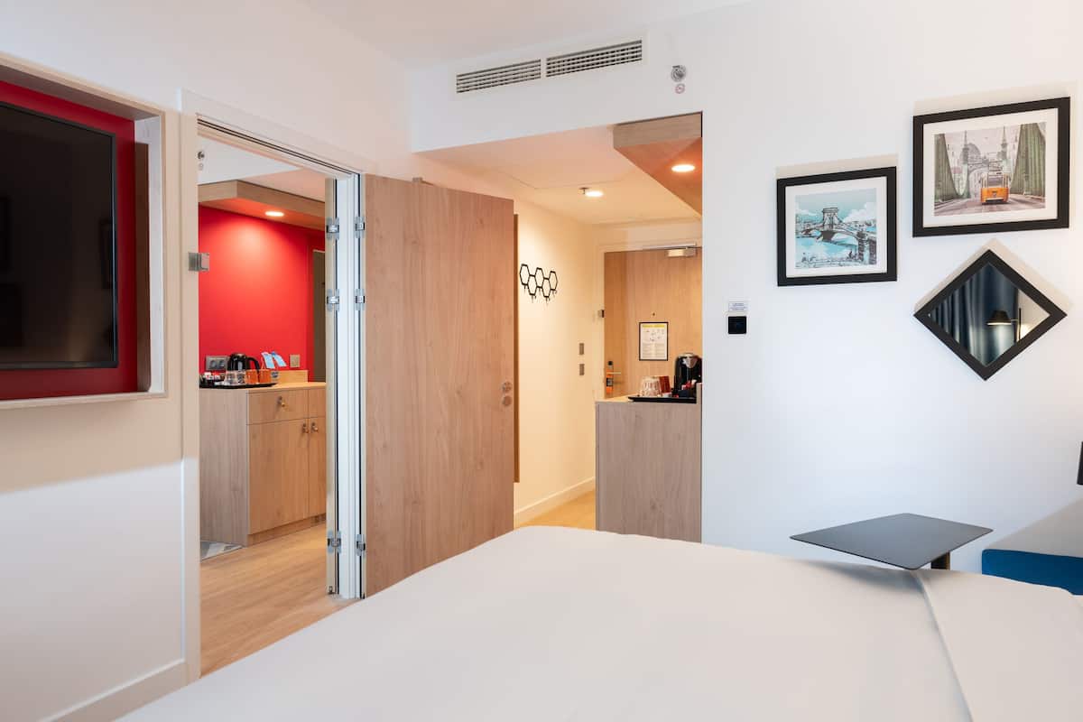 A modern hotel room with a bed, a desk, and a kitchenette. There is a door leading to another room and a wall with framed pictures.