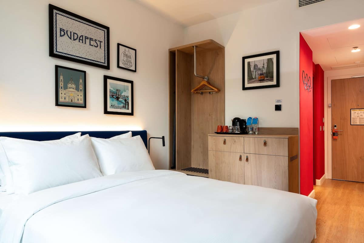 A hotel room with a bed, a wooden wardrobe, a coat rack, and framed pictures on the wall.