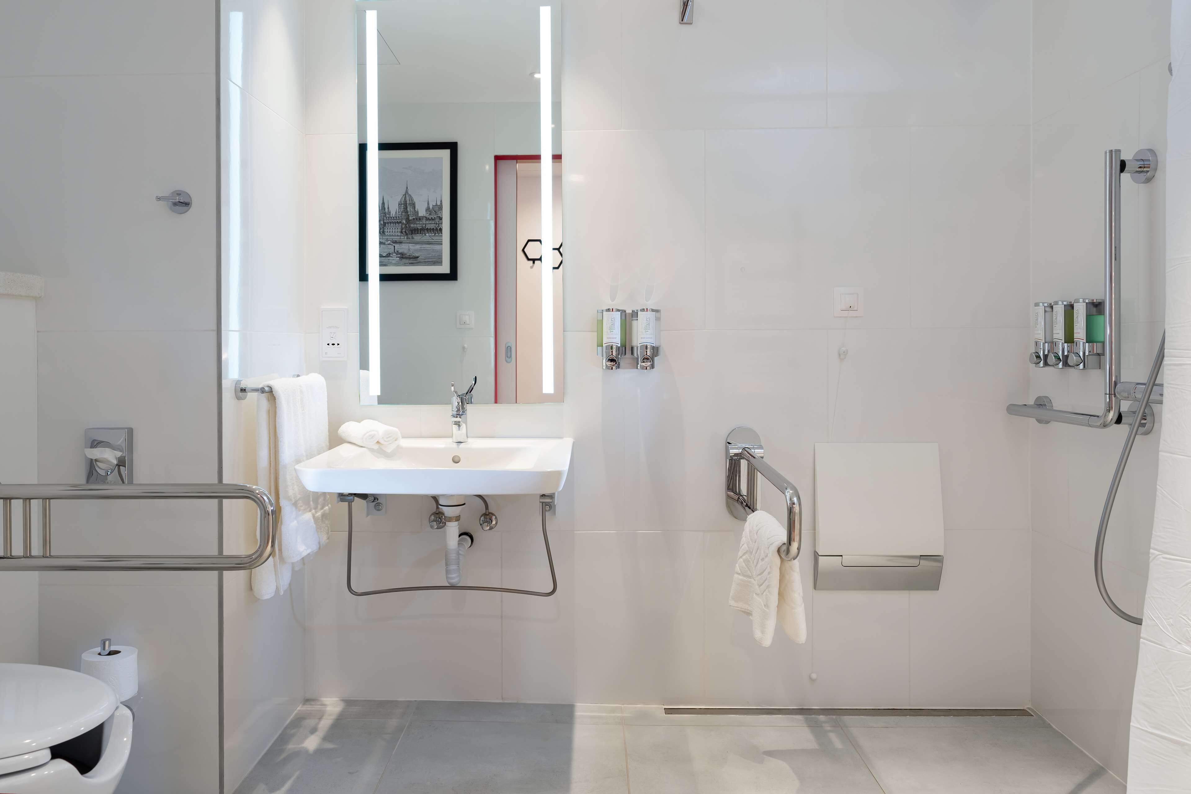 A modern bathroom with a white sink, mirror, and shower. There are handrails and a towel rack.