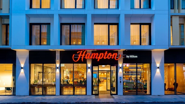 Un hotel con fachada moderna, amplias ventanas y un llamativo letrero de 'Hampton by Hilton'.