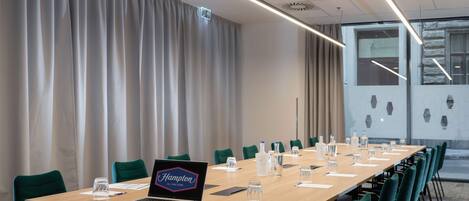 Un salón de conferencias con una larga mesa, sillas verdes, una computadora portátil, botellas de agua y un cartel que dice "Hampton by Hilton".