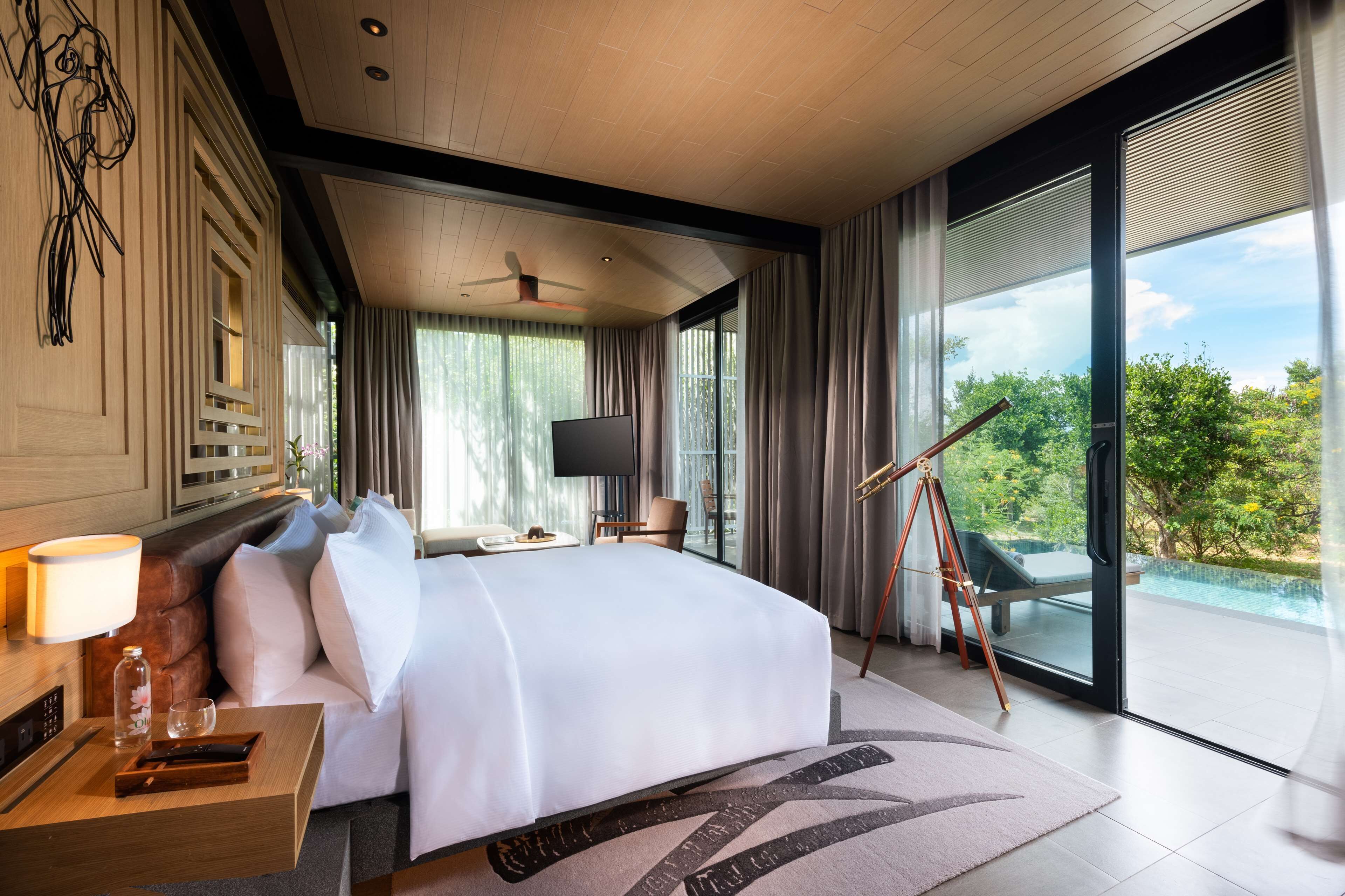Una habitación de hotel moderna con una cama grande, un escritorio con lámpara, televisión y un balcón con vistas al verdor.