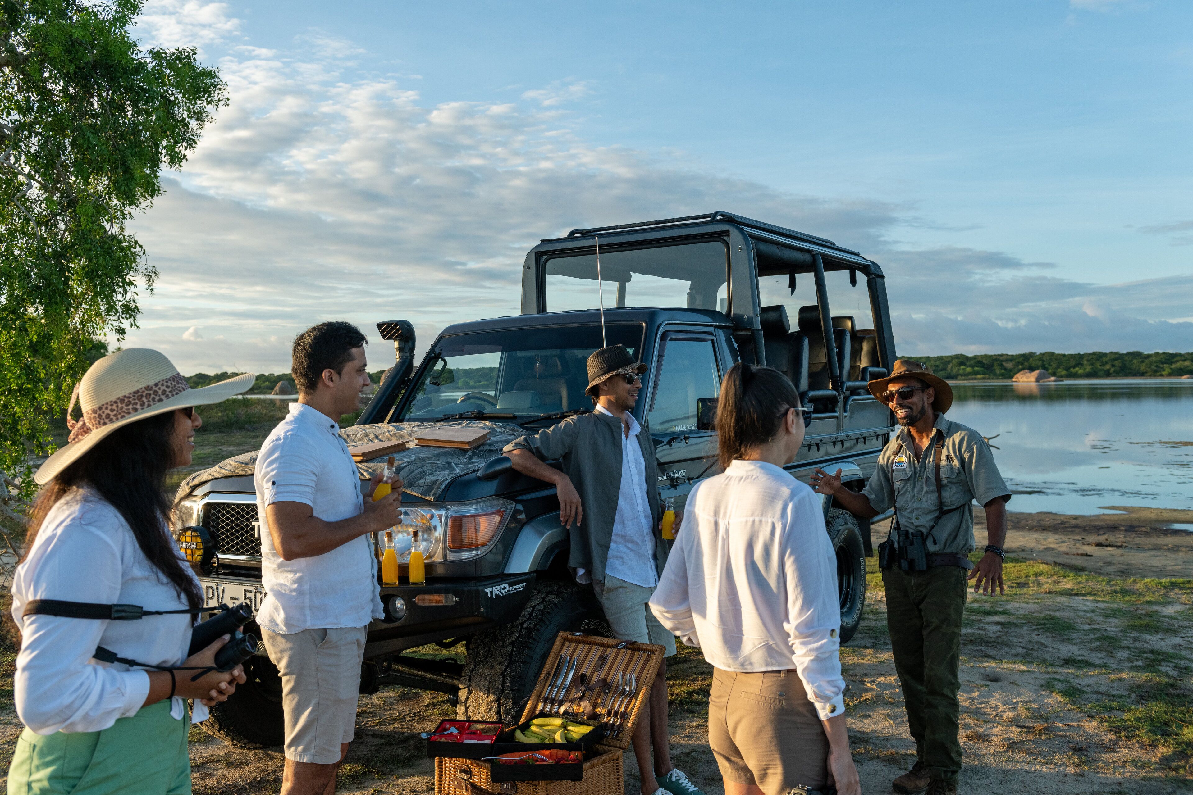 Un grupo de personas cerca de un SUV negro, con una instalación para picnic que incluye una cesta con comida y bebidas, junto a un cuerpo de agua.