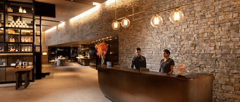 A modern hotel lobby with a curved reception desk, a stone wall, and a bar area with shelves of bottles.