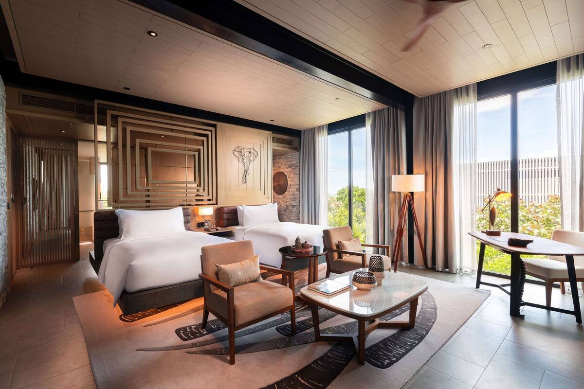 A hotel room with two beds, a sitting area with a table and chairs, a large window with curtains, and a view of greenery outside.