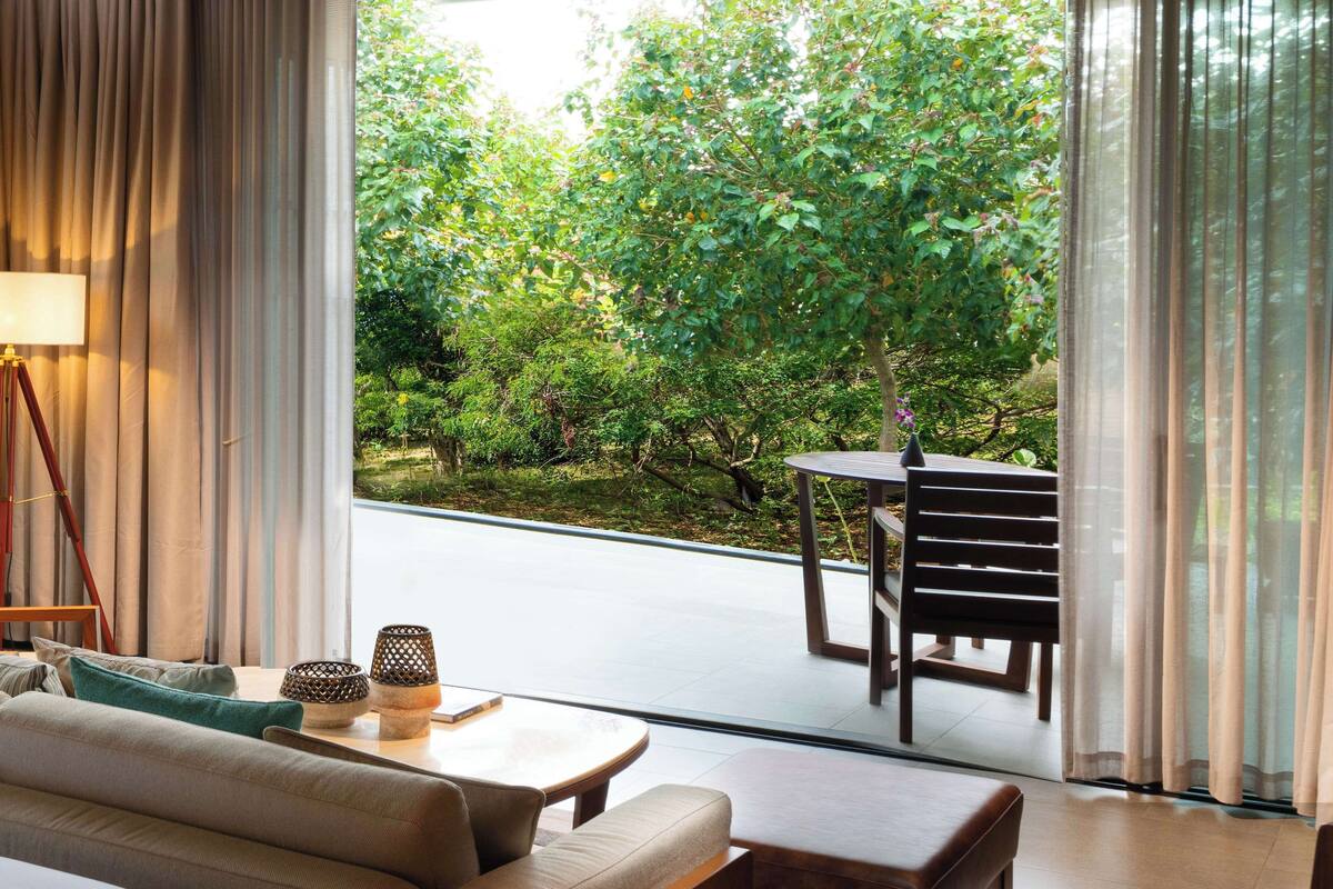 A modern living room with a sofa, a small table, and a chair, overlooking a garden with trees and a pathway.