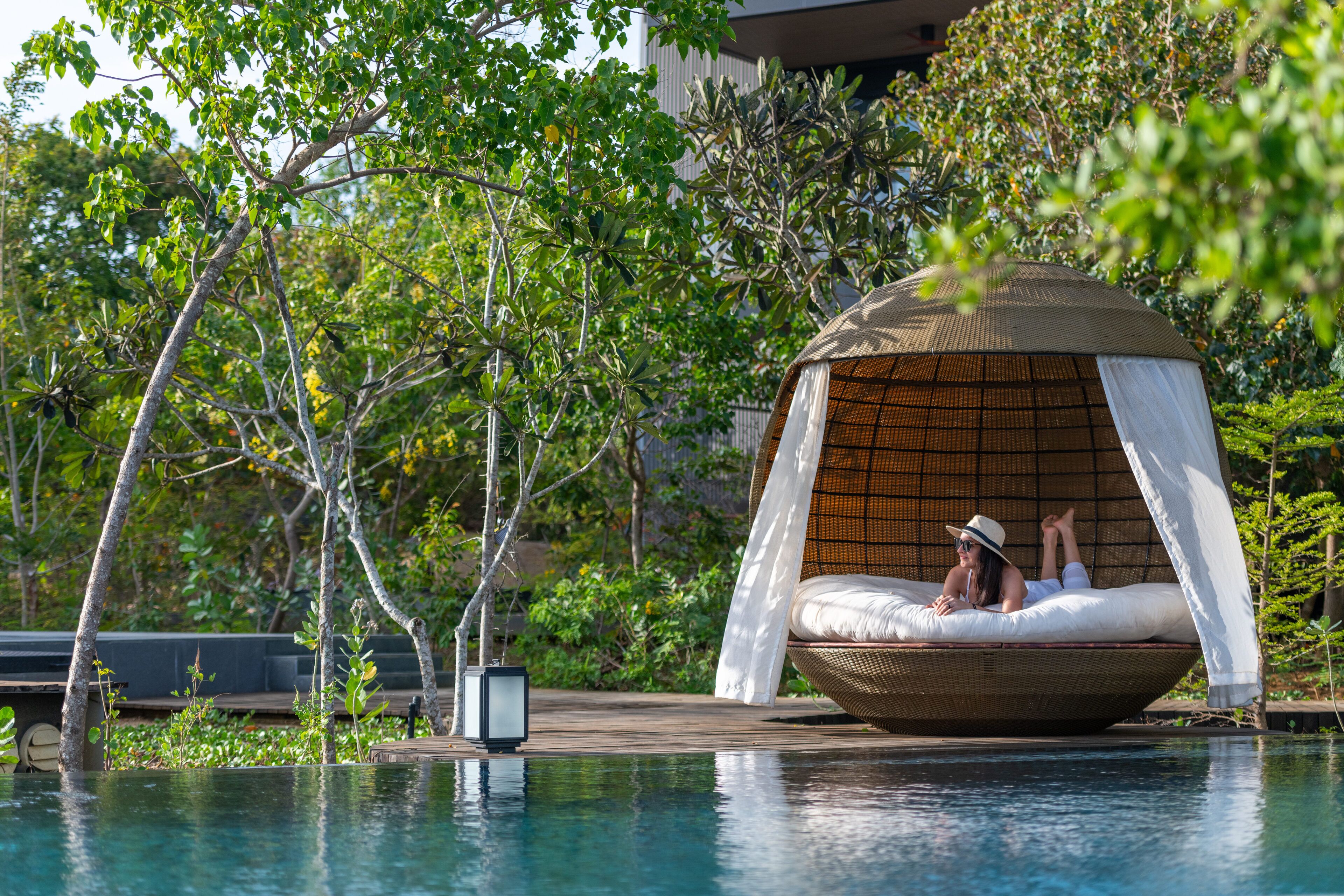 A secluded outdoor seating area with a wicker chair and canopy, surrounded by lush greenery and a pool.