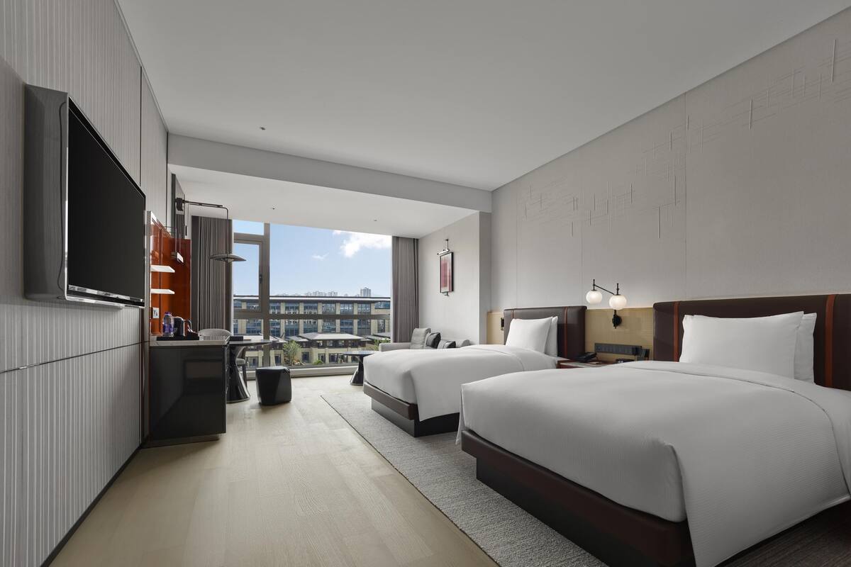 A hotel room with two beds, a flat-screen TV, a desk with a chair, a small trash bin, and a view of the cityscape.