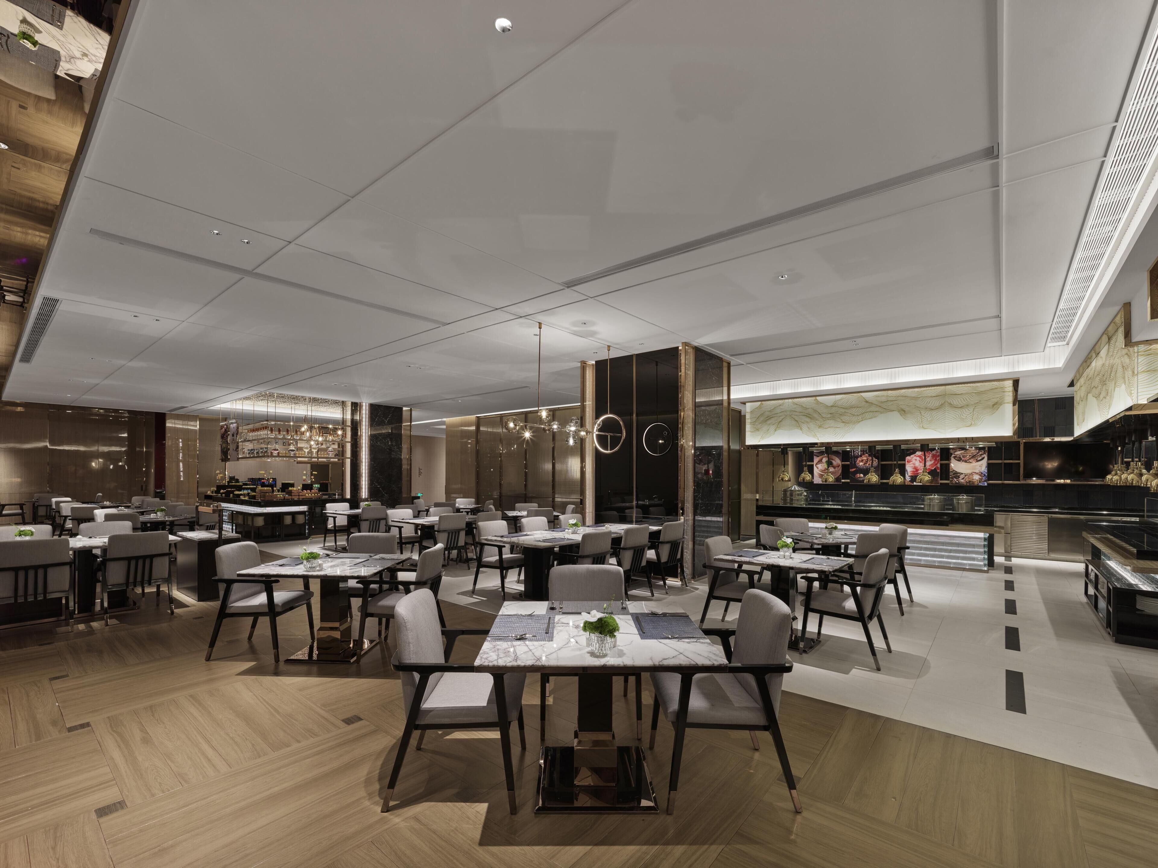 A modern restaurant with multiple dining tables and chairs, a bar area, and a high ceiling with recessed lighting.