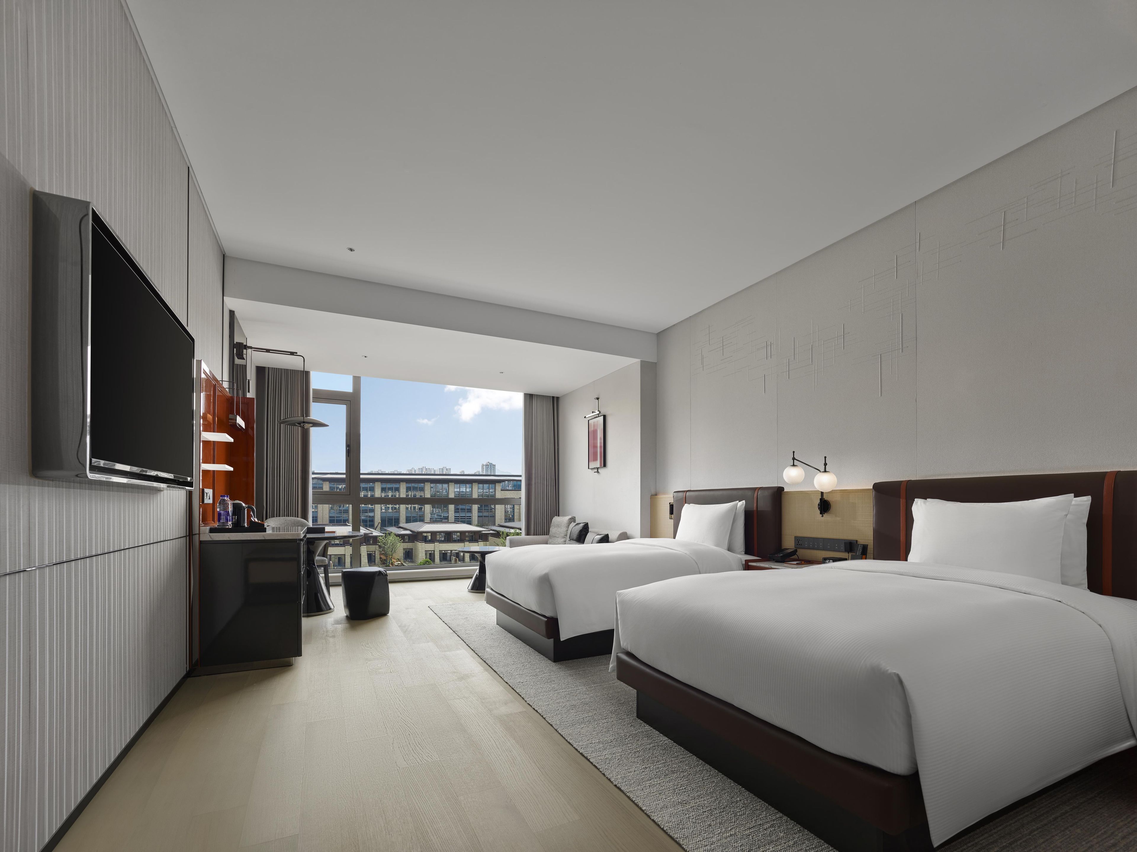 Een hotelkamer met twee bedden, een flatscreen televisie, een bureau met stoel, en een groot raam met uitzicht op de stad.