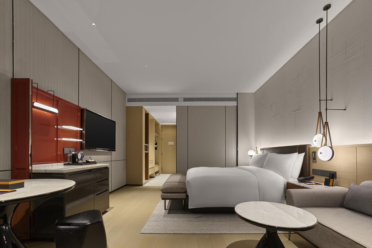 A modern hotel room with a bed, bedside table, a desk with a chair, a television, and a kitchenette.