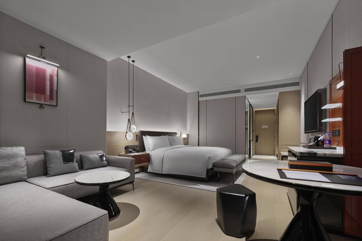 A modern hotel room with a sofa, a round table, a bed, a TV, and a bathroom.