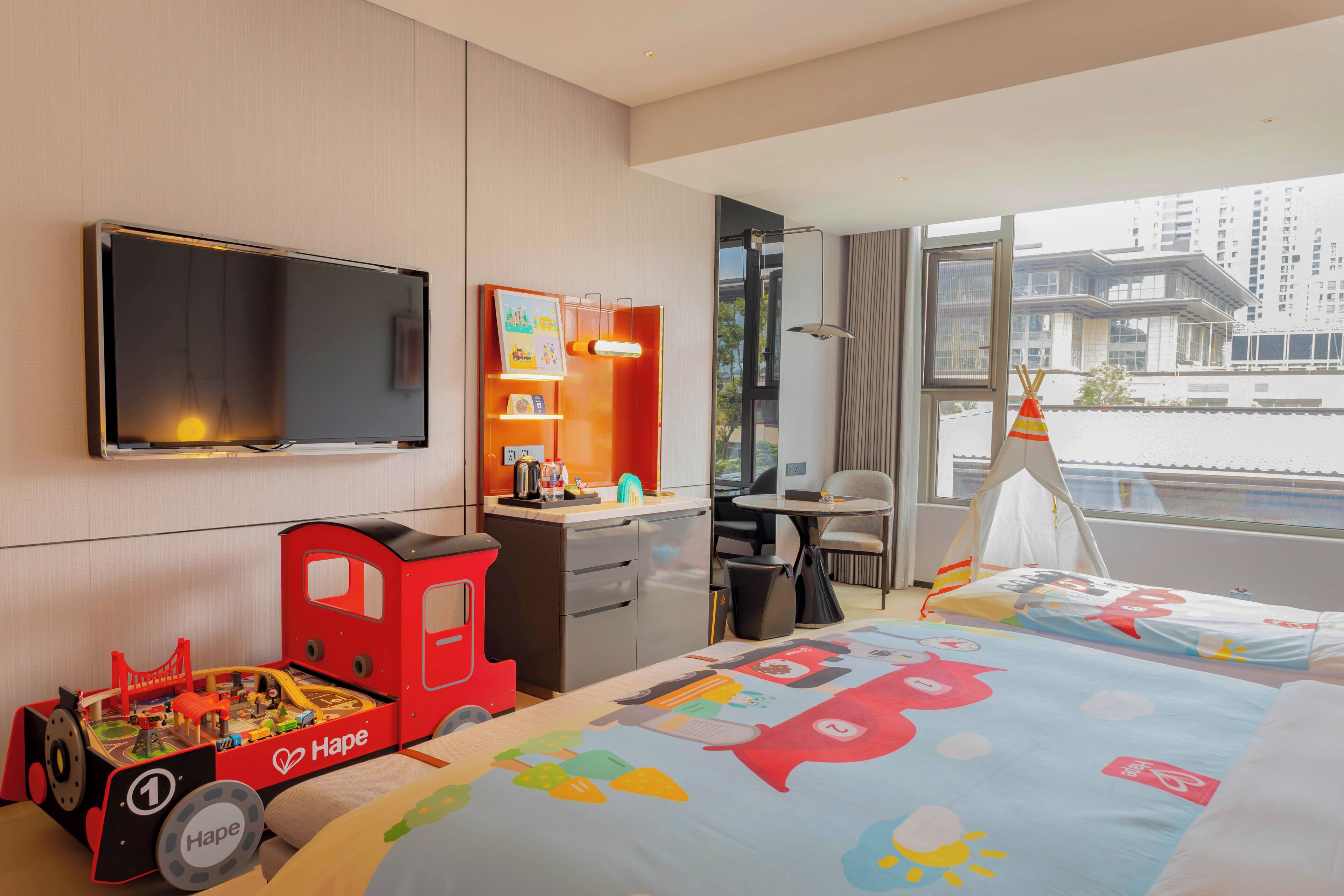 A hotel room with a bed, a desk, a television, a toy train, and a window with a view of buildings.
