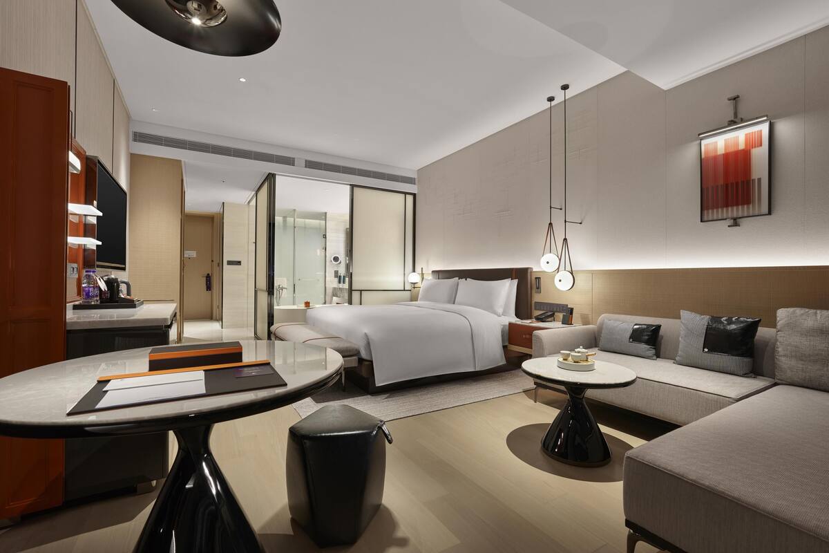A modern hotel room with a bed, sofa, and a small table. There is a mirror, a wall-mounted light, and a ceiling light.