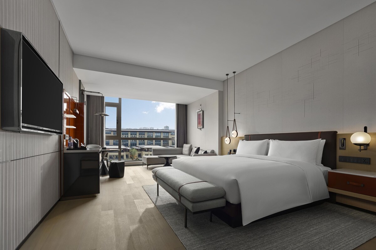 A modern hotel room with a large bed, a desk, a flat-screen TV, and a city view.