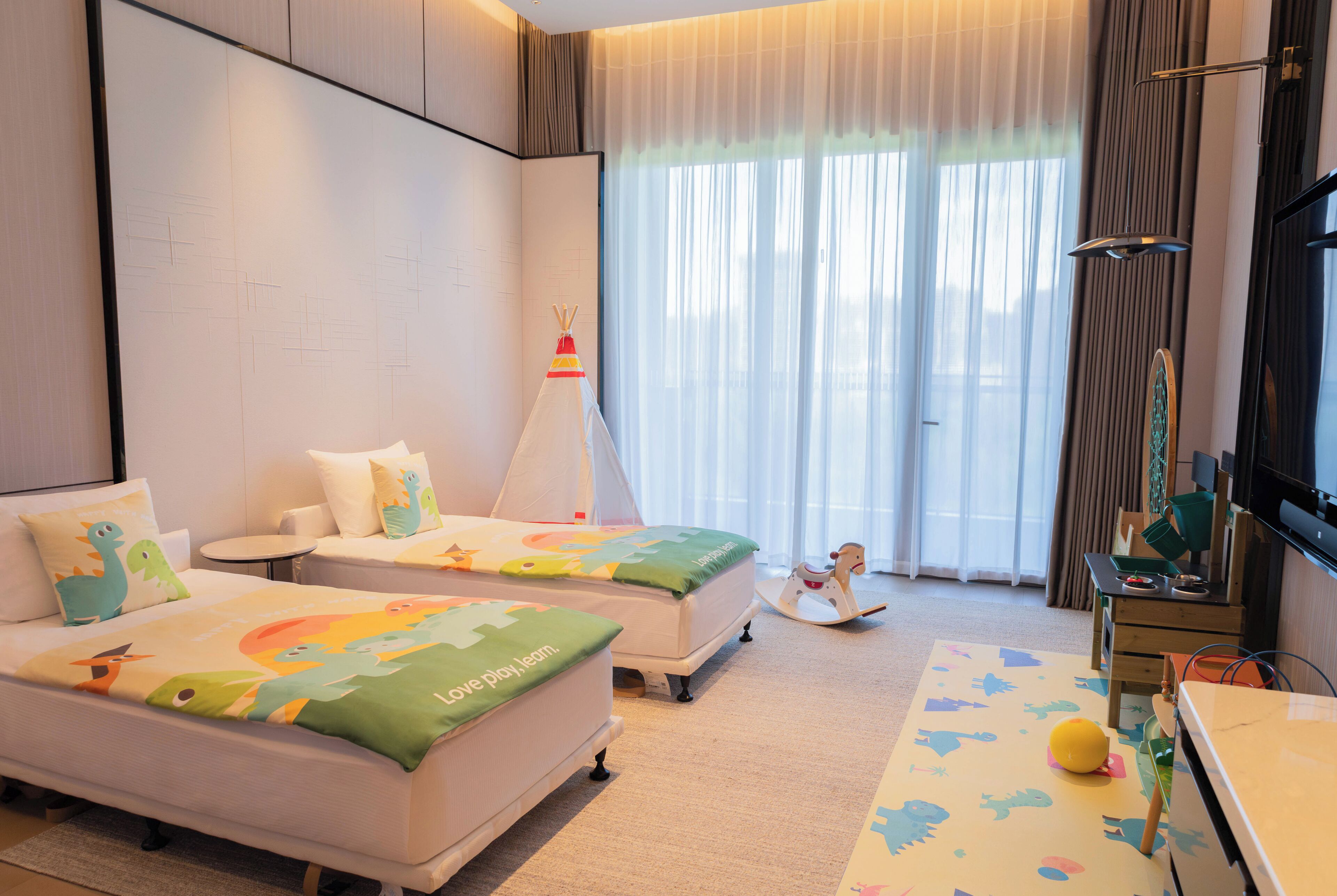 A hotel room with two beds, a desk, a chair, a TV, and a large window with curtains.