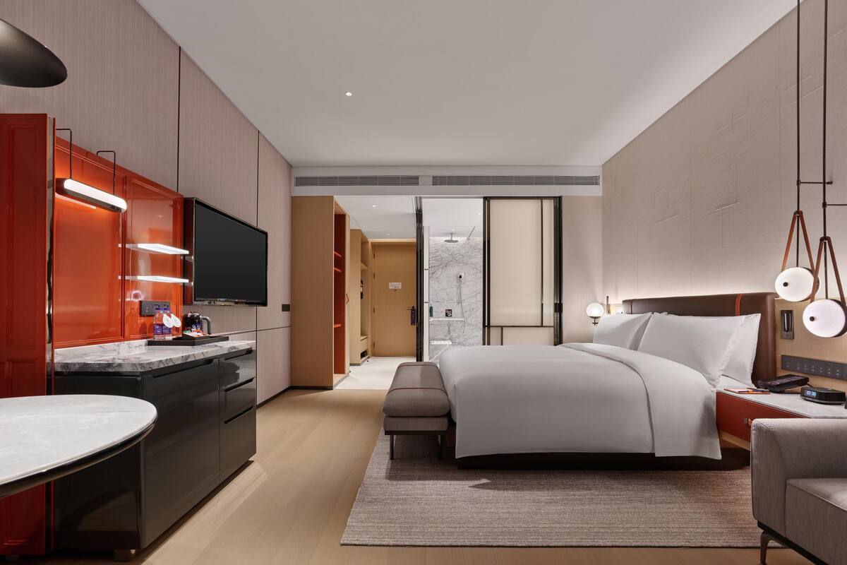 A modern hotel room with a large bed, a desk with a flat-screen TV, a built-in kitchen with a marble countertop, and a sliding door leading to a bathroom.