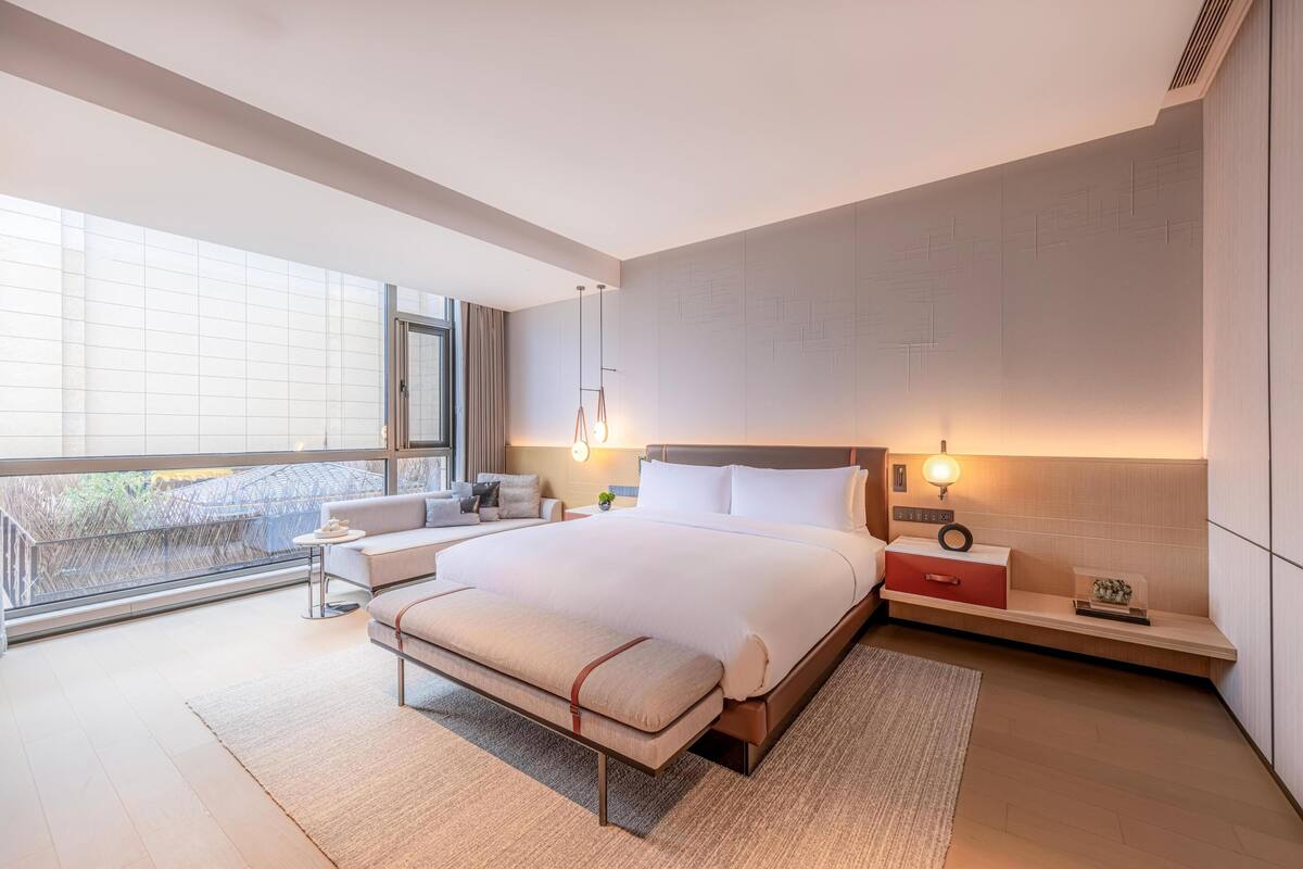 A modern bedroom with a large bed, bedside tables, a bench, and a view through a large window.