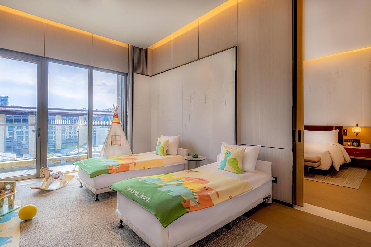 A hotel room with two beds, a large window, a view of the city, and a children's play area.