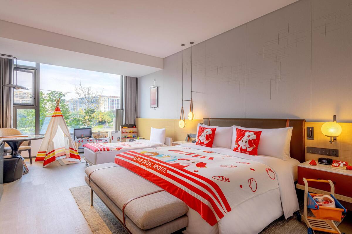 A hotel room with a large bed, a desk, a chair, a small table, a bench, a TV, a lamp, and a window with a view of buildings and trees.