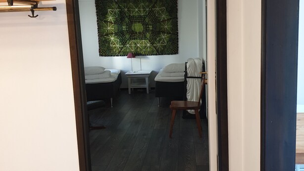 Ein Zimmer mit zwei Betten, Holzboden, einer an der Wand hängenden grünen Wandteppich und einem kleinen Tisch mit Lampe.