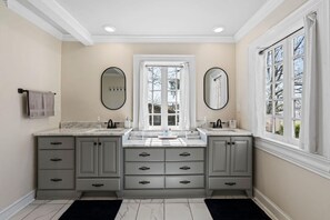 A bathroom with two sinks, grey cabinets, a large mirror, and a window with curtains.