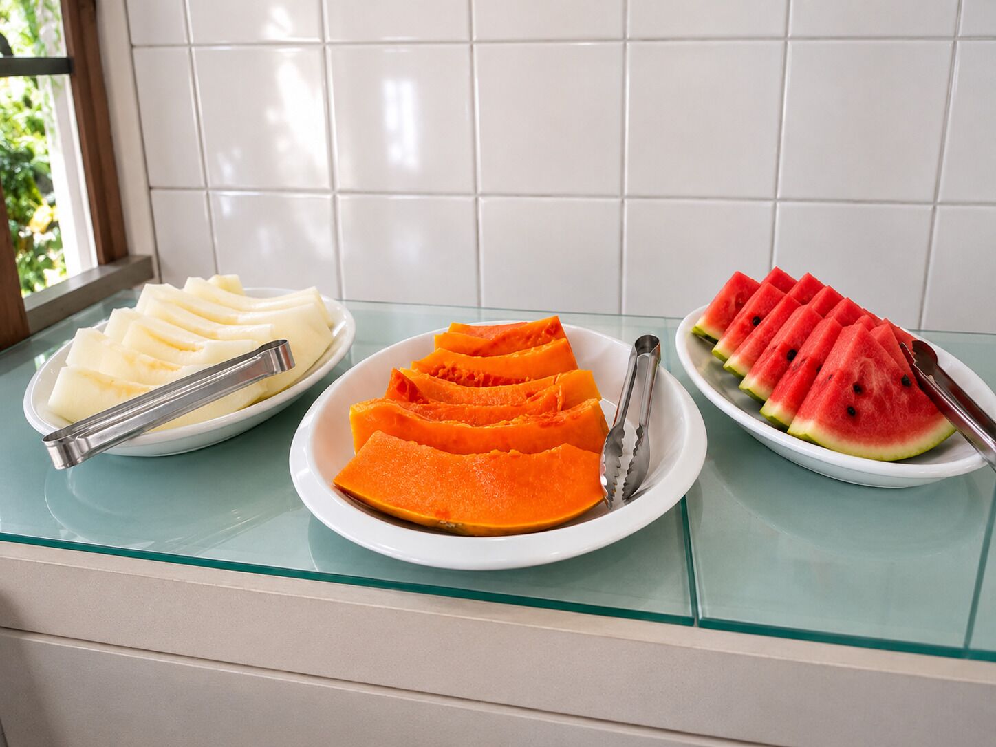Un comptoir de cuisine sur lequel se trouvent trois assiettes de nourriture : des tranches de fruits blancs, des tranches d’orange et de la pastèque.